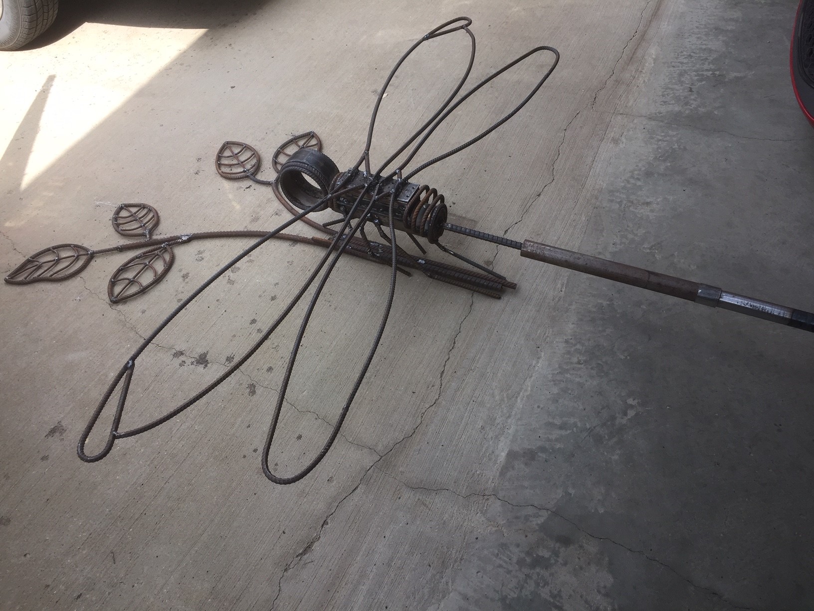 A dragonfly torso, wings and head sculpted out of metal, attached to a pole with five leaves at the top.